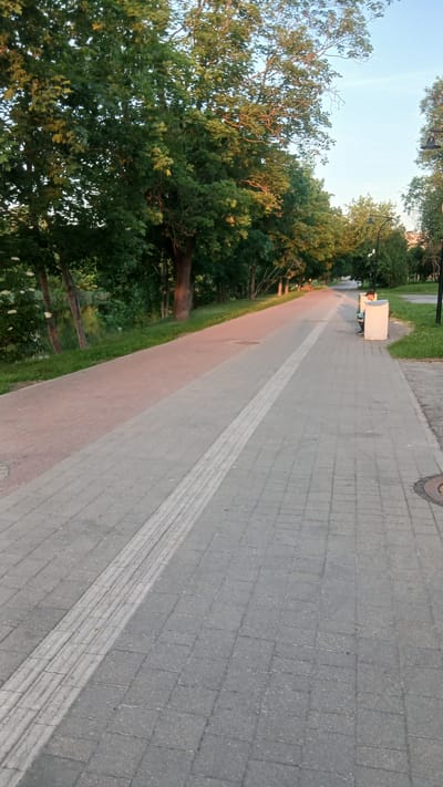 The image shows a paved pathway in a park or along a body of water, with mature trees lining the left side. The pathway is made of gray interlocking pavers, with a tactile paving strip running along its center. To the left of the pathway, there is a grassy area and then a body of water with trees and vegetation reflected in it. The sun appears to be setting or rising, casting a warm light on the trees and pathway. On the right side of the pathway, there are streetlights and a park bench with a person sitting on it. The overall impression is of a tranquil, well-maintained public space. The location is identified as Jelgava, Latvia.