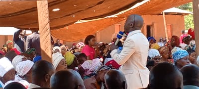 The image shows a large gathering of people, mostly women, seated outdoors under a makeshift canopy of brown fabric in Kalemie, Democratic Republic of the Congo. The majority of the women are wearing traditional headwraps in various colors and patterns.
A man in a light beige suit is standing and speaking into a blue microphone while holding a bottle of water. He appears to be addressing the crowd. Another woman wearing a pink shirt is holding a cell phone.
Behind the crowd, there is a mud-brick building with small windows. A few individuals are standing near the entrance of the building. The atmosphere appears to be one of a community event or gathering.