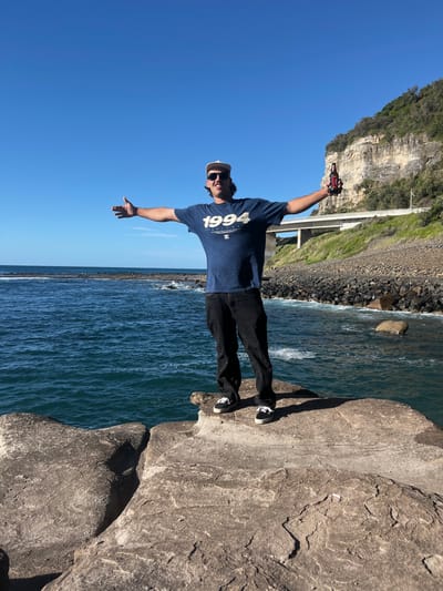 A single male individual stands centrally on a flat, light grey rock formation at the ocean's edge in Coalcliff, Australia, in the vicinity of the Seacliff Bridge. The man faces the viewer, his arms extended horizontally. He wears a light-colored baseball cap, dark sunglasses, a dark blue t-shirt bearing "1994" and "DC SHOES CO." branding, dark jeans, and black and white lace-up sneakers. In his right hand, he holds a dark glass bottle with a red label. The ocean, deep blue and exhibiting minor surface chop, extends across the left and central background. To the right, a rocky shore composed of dark pebbles and boulders borders the base of the Illawarra escarpment, a steep, light-colored sandstone cliff with green vegetation atop and along its face. A section of the concrete Seacliff Bridge, identifiable by its piers and guardrail, is visible spanning above the rocky shore and against the cliff face. The sky is clear and bright blue. A discarded white object, consistent with a cigarette butt, is present on the rock platform in the lower right foreground.