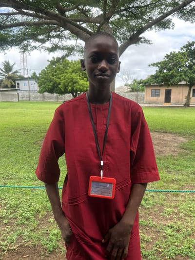 A young boy, appearing to be of Nigerian ethnicity and possibly under 18 years of age, stands outdoors in Itoga, Nigeria. He is wearing a traditional red garment, possibly an agbada, with a black lanyard around his neck. Attached to the lanyard is a bright orange identification badge holder containing a white card with handwritten text in black ink, some of which is legible. The boy's hands are tucked into the front pockets of his garment, and he is looking directly at the camera with a neutral expression. He has short hair and dark skin.
The background consists of a grassy field, a mature tree with an expansive canopy, and in the distance, buildings and a cell tower. The sky is overcast. The scene suggests a casual outdoor setting, potentially within a school or community compound, given the presence of the ID badge.