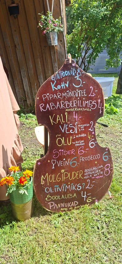 A dark brown, violin-shaped wooden chalkboard menu is displayed outdoors on a grassy area, leaning against a wooden wall structure. The chalkboard lists various food and drink items with prices written in different colored chalk. Items include "GINTOONIK" (7.00), "KOHV" (3.00), "PIPARMÜNDITEE" (2.00), "RABARBERILIMPS" (4.00), "KALI" (4.00), "VESI" (2.00), "ÕLU" (4.00), "SIIDER" (6.00), "VEIN" (6.00), "PROSECCO" (6.00), "MULGIPUDER" (8.00), "OLIVIMIIKS?" (2.00), "SOOLAJUBA" (4.00), and "PANNUKAD" (6.00). Some items have additional descriptions like "AUGUDEGA VÕI ILMA" (with or without holes), "ALKOHÖVABA" (alcohol-free), "ALE, GOH PREMIUM" (ale, coffee premium), "SOOLALIIHA HAPUKURK KÜLM KASTE VÕI VEGAN" (cured meat, pickles, cold sauce or vegan), and "MOSSI VÕI MARJAD" (moss or berries). A pot of bright orange and red marigolds sits in the lower left corner. The background shows a wooden wall, green foliage from trees, and a glimpse of a white bin. The overall setting suggests an outdoor market or cafe. The location is the city of Viljandi, Estonia.