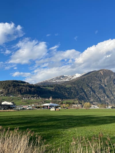 The image presents a landscape in Sterzing-Vipiteno, Italy, with a mountain range partially covered in snow as a prominent backdrop. The sky features a mix of blue and white clouds. In the foreground, there is a field of green grass with reeds along the edge, near the horizon. A partially visible building with a curved roof, possibly a sports facility, is positioned along the horizon line. Buildings in the city are visible.