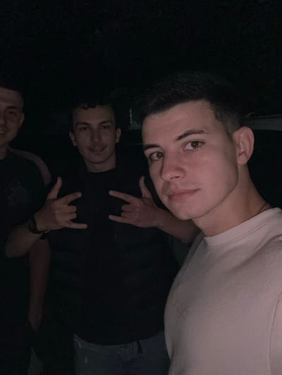 A group of three young men is captured in a dark, low-light setting, likely at night in Sevlievo, Bulgaria. The image is oriented sideways. The most prominent figure, a young man with short, dark curly hair, fair skin, and brown eyes, is positioned at the top. He wears a light-colored, possibly white or light beige, crew-neck t-shirt and a thin chain around his neck. On the t-shirt, the embroidered brand logo "H&M" is visible. He looks towards the camera with a neutral to slightly smiling expression. Below him, partially obscured by darkness, is a second young man with dark hair, wearing a dark t-shirt. His right hand is visible, making the "rock on" hand gesture. Further down, only the top of a third young man's head is visible. He also wears a dark t-shirt, and his left hand is seen making the "rock on" gesture, with a watch on his wrist. The background is completely dark, providing no additional environmental cues. The overall mood appears casual and friendly, suggesting a social gathering.