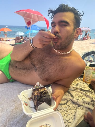 In Badalona, Spain, a man is lying on a towel on a crowded beach, holding a piece of chocolate cake. He is wearing green swim trunks and a shell necklace, and a bracelet on his wrist. The ocean and several beach umbrellas are visible in the background. He is bringing a bite of cake to his mouth.
