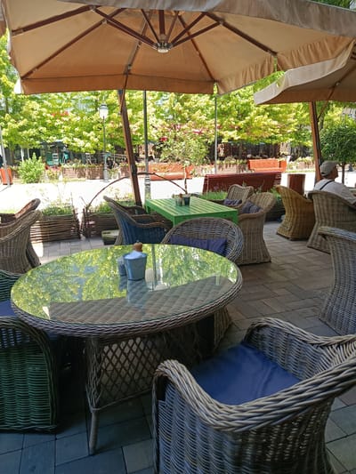 The image depicts an outdoor seating area of a cafe or restaurant, likely in the city of Grozny, Russia, given the context provided. In the foreground, a round glass-topped table made of woven material sits on a patterned paved surface. Several chairs, also of woven material with dark blue cushions, surround the table. One chair is partially visible on the left, and another is prominent in the lower right corner.

Behind the table, there is a green rectangular table with chairs around it. Further back, several other woven chairs and tables are visible. A man wearing a light-colored cap and a white shirt is seated in one of the chairs, facing away from the camera. In the background, lush green trees with some red foliage create a backdrop. There are also wooden planters and a lamppost. The scene is covered by large, light-colored umbrellas, suggesting a sunny day. The overall impression is a pleasant, shaded outdoor dining environment.