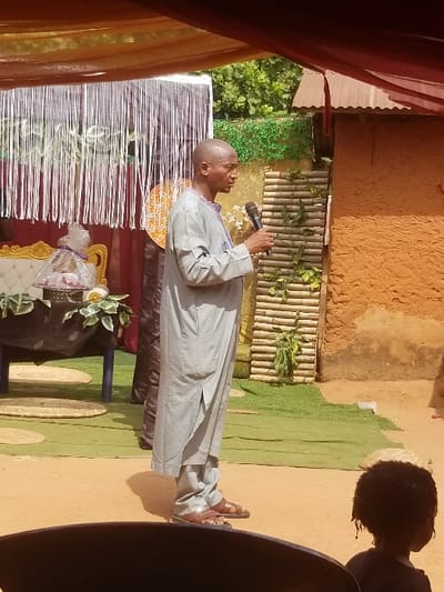 The image presents an outdoor scene in Ochadam, Nigeria, under a temporary canopy composed of draped orange/gold and maroon fabric. An adult male, bald and light-skinned, stands in profile facing right, wearing a light grey tunic, matching trousers, and brown leather sandals. He holds a black microphone to his mouth, appearing to speak.
To the left and slightly behind the speaker, a decorated area features a white-framed, upholstered chair with gold accents. A dark-covered table positioned in front of this chair holds a cellophane-wrapped gift basket adorned with a purple ribbon and green foliage. Behind these items, a backdrop of vertical silver strips is visible against a maroon fabric.
The background includes distant green foliage and trees. To the right of the speaker, a fence constructed from horizontal bamboo slats is visible, decorated with artificial green vines. Behind this fence and slightly right, a reddish-brown, textured mud-built structure with a corrugated metal roof is present. A patch of green artificial turf or fabric lies on the unpaved earth ground in the mid-right. In the lower right corner, the back of a small child's head with dark, curly hair is partially visible, while a dark, curved object occupies the immediate foreground in the lower left.