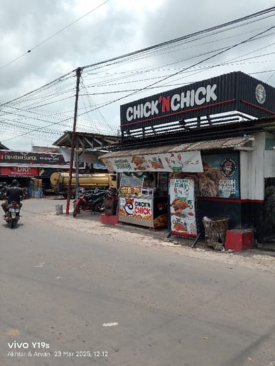 The image shows a street scene in Kecamatan Haurgeulis, Indonesia. A small food stall with a sign that reads "Chick 'n Chick" in red and white letters is prominently featured. A person riding a motorcycle is driving down the street. The motorcycle is parked near a truck and a stand with several motorcycles parked. There are power lines overhead, and the sky is partially clouded. There are several colorful signs depicting fried chicken and other food items. The stall's walls are black and adorned with numerous additional signs. The stall's roof is made of corrugated metal. The pavement is gray and weathered. There are some plants and a trash bin outside the stall.