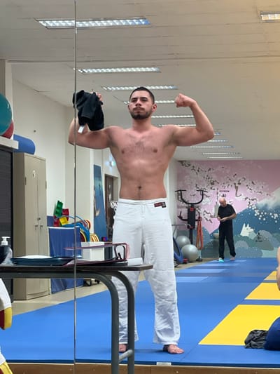 A shirtless, muscular man in white pants poses flexing in front of a mirror. He holds a black cloth in one hand. The reflection shows a gym with blue and yellow mats, exercise equipment, and another person in the background. The walls are light-colored, and one wall has a decorative cherry blossom design. The man's reflection is visible, suggesting a gym or studio setting.