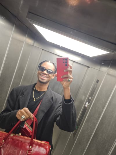 A person is taking a selfie in an elevator. The person is wearing sunglasses, a black jacket, and gold jewelry. They are holding a red smartphone in their right hand. In their left hand, they are holding a red leather briefcase. The elevator has metal walls and a fluorescent light. The location is Pantin, France.