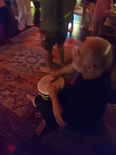 A man with a shaved head and wearing a black shirt is seated and playing a djembe drum. He is sitting on a gray cushioned surface. His bare feet are visible. A patterned rug covers the floor. Other people are standing and milling around in the background. Their clothing is varied, including pants and shorts. The location is possibly indoors, based on the lighting and rug. There is no visual information to identify Lokaserena, Indonesia.