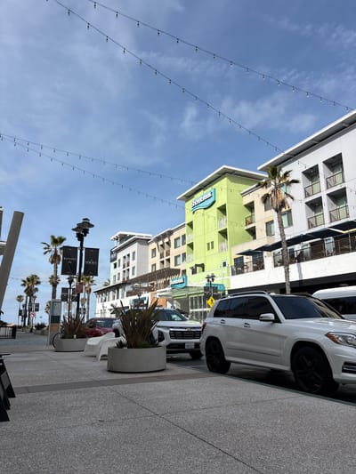 This image presents a rotated, upward-angled view of a modern street scene in Huntington Beach, California, under a clear blue sky with scattered wispy clouds. The time of day is bright daylight.
In the foreground, a light gray, speckled sidewalk stretches alongside a street where vehicles are parked. A white SUV, appearing to be a Mercedes-Benz GL-Class with black wheels, is prominent, and behind it, a California license plate reading "9YO9021" is visible on another car. Concrete planters containing spiky, ornamental plants and a white outdoor chair are placed along the sidewalk.
Overhead, multiple parallel strings of lights extend across the expansive blue sky. Palm trees, common in coastal California, punctuate the scene, standing tall between the buildings.
The architecture consists of multi-story, contemporary buildings. A distinctive light green building with a prominent blue "Shorebreak" logo suggests a hotel or resort, a detail reinforced by a smaller "hotel" sign below it. Other buildings feature gray, white, and beige facades with numerous windows and balconies. Further down the street, black banners hanging from lampposts display "WELCOME TO" and "SHOREBREAK". A yellow diamond-shaped sign depicting a person on crutches is also visible, indicating a pedestrian area or accessibility notice.
The scene captures a vibrant and modern urban environment, typical of a coastal resort town. There are no people visible, but the setup suggests a well-maintained commercial and pedestrian zone.