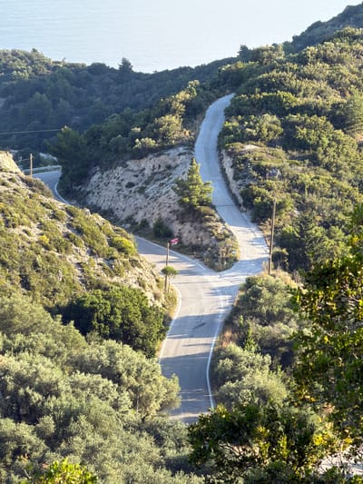 The image depicts a winding road cutting through a densely vegetated, hilly landscape, presumably located in the Municipal Unit of Sfakiotes, Greece. The road is paved with a light gray asphalt and has white line markings on the edges. The terrain is covered in lush green vegetation, including trees and shrubs. There are utility poles running along the road. In the background is the ocean.