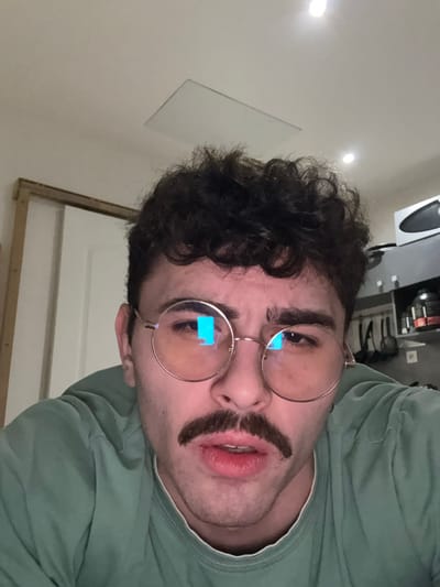A close-up, rotated image features a man with dark, curly hair and a fake mustache. He wears round, gold-rimmed glasses with blue reflections. His expression appears neutral. The background includes a plain white wall and a hint of a white door. The man is wearing a light green shirt. A framed white object is barely visible on the left side of the image, near the wall.