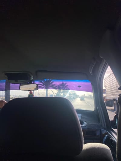 The scene is observed from inside a vehicle, positioned in a rear passenger seat, looking forward through the windshield onto a highway in La Cruz del Pastel, Venezuela. The vehicle's dark grey headliner occupies the upper portion of the frame. A dark grey driver's seat headrest is prominent in the lower center foreground. A rearview mirror hangs centrally, with a small red and white decorative object suspended from it. The upper windshield features a wide, iridescent purple-blue tint band, to which a small dark, looped object is adhered on the right side. The driver's left hand is visible on the steering wheel on the far left.

Beyond the windshield, a multi-lane paved road extends under direct, bright sunlight, indicative of a morning or late afternoon setting. Numerous palm trees line the roadside, silhouetted against the bright sky and the windshield's tint. The distant landscape reveals arid terrain with low-lying hills under a clear sky. Other vehicles are present on the road: a light-colored sedan is visible in the distance, and the right side of a large white semi-truck occupies an adjacent lane to the vehicle's immediate right. Portions of the vehicle's dashboard, including air vents and a textured mat, are visible beneath the windshield.