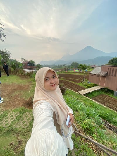 A young woman in a tan hijab and white long-sleeved shirt is standing outdoors, holding a white rectangular object with a gray cord attached. She is smiling at the camera. Behind her, there are tiered rice paddies, a brown wooden building, and a mountain in the distance shrouded in mist. Tents are set up in the rice paddies. In the foreground, the ground is covered in green grass and soil. Two figures are visible in the lower part of the image. Based on visual cues, this image was most likely taken in Tamiajeng, Indonesia.