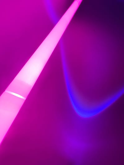 This image, captured in Derby, United Kingdom, presents an abstract scene dominated by artificial light and color. A bright, elongated object, likely a light tube, emits a strong pink-magenta glow and features a distinct horizontal white band. This object is positioned diagonally, extending from the upper-left corner towards the lower-right. The immediate background and surrounding environment are illuminated in shades of deep magenta and purple. In the lower-right quadrant, a curved arc of blue-violet light is cast upon a surface, exhibiting a subtle texture. No individuals, specific objects beyond the light sources, or discernible actions or interactions are present in the scene.