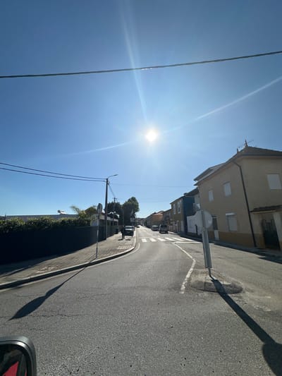 The image depicts a street scene in Gondomar, Portugal, during a bright, sunny day. The street is asphalt, with a concrete curb and sidewalk on the right. Buildings line the left side of the street, varying in height and color, but predominantly beige and tan. A traffic sign is visible on the right side of the road. Power lines run horizontally across the upper portion of the frame, intersecting with the sun's glare. A few parked cars can be seen further down the street, and some sparse vegetation is present on the side of the road. The camera seems to be positioned inside a vehicle, as indicated by a partial view of a side mirror in the lower right corner. The shadow of a light pole is visible stretching across the road.