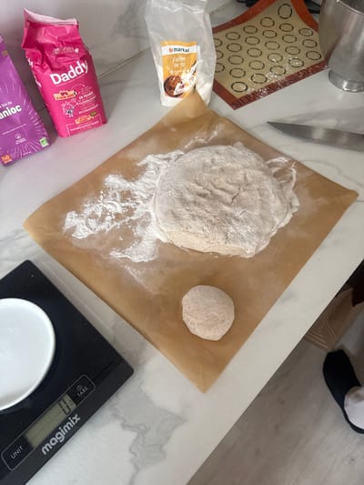 This is a top-down view of a kitchen counter, displaying ingredients and tools associated with baking. A larger mound and a smaller ball of what appears to be dough sit on a square piece of parchment paper. Scattered flour surrounds the larger mound.
Adjacent to the parchment paper, a package of "markal Farine de riz" (rice flour) is visible, along with a pink bag labeled "Daddy" with French text and imagery, and a purple package with the word "anioc". To the left of the dough, a silicone baking mat with circular imprints sits next to a silver knife and a stainless steel mixing bowl. To the right of the parchment paper, there is a black "magimix" kitchen scale with a white dish.
A foot wearing a sock and a black slipper appears in the upper right corner of the image. The counter is made of marble.
Based on the packaging of "Daddy" which states "Cultivées en France" (Grown in France) and "Conditionné en Champagne" (Packaged in Champagne), this image was most likely taken in France. The cooperative origin "Cooperative Agricole Francaise" of the "Daddy" product further narrows the origin. It's unlikely that the image was taken in the city of Levallois-Perret, France.