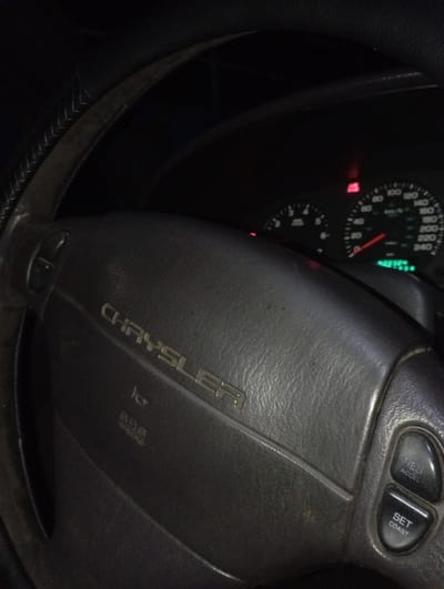 An interior shot of a vehicle, taken from the driver's perspective in Kurganinsk, Russia, depicts a dark environment with the instrument cluster illuminated. The central subject is a black Chrysler steering wheel, featuring the "CHRYSLER" emblem embossed in the center. Below the emblem, "SRS AIRBAG" is visible, along with a horn symbol. The right spoke of the steering wheel contains two control buttons: "RES ACCEL" and "SET COAST." The steering wheel rim, partially visible on the left, exhibits a stitched texture. In the background, the vehicle's illuminated dashboard instrument cluster is visible, displaying a speedometer marked up to 240 km/h with its needle at zero, and a tachometer marked up to 8 (x1000 RPM) also at zero. A red warning light is illuminated above the speedometer, and another red warning light is illuminated to the left of the tachometer. A green digital display below the speedometer shows "48754" and additional obscured green digits. No individuals are visible in the frame.