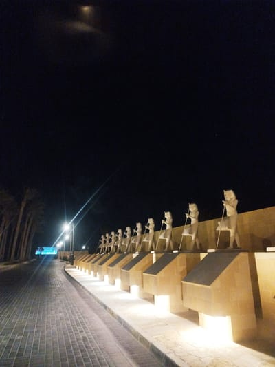 The image displays a nighttime scene featuring a paved pathway and a row of illuminated ancient Egyptian-style statues. On the right side of the frame, a long line of uniformly spaced, light-colored statues depicts figures holding staffs or scepters. These statues stand on a raised, light-brown colored wall or pedestal, which is accented by integrated uplights that cast bright illumination onto the figures and the wall's base.
To the left, a wide, tiled or paved walkway curves into the distance. This path is bordered by several streetlights that emit strong beams of light, creating distinct lens flares against the dark night sky. Tall, slender trees, possibly palm trees, are silhouetted along the far left edge of the pathway. In the far distance, at the curve of the path, a structure glows with a distinct blue light.
The sky is dark blue-black, indicative of nighttime, with a faint, blurry light source visible near the top center. The primary light in the scene comes from artificial sources, including the streetlights and the integrated uplights on the statue display, which create a dramatic and well-lit corridor. No individuals are visible, and no specific activity is taking place. The scene appears to be an outdoor cultural or historical display. There is no visible text in the image.