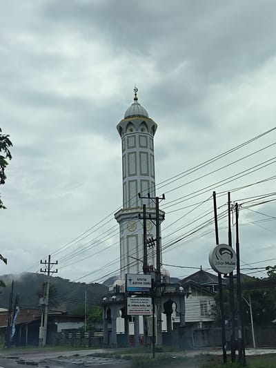 This image captures a street view in Lampisang, Indonesia, dominated by a tall, ornate mosque minaret piercing a cloudy sky. The minaret is white with intricate gold detailing and topped with a crescent moon and star. In the foreground, a web of power lines crisscrosses the frame, supported by utility poles.

Below the minaret, an arched entrance with Islamic architectural motifs leads to a building. To the left of the entrance, a sign for "APOTEK KARTINI FARMA" is visible, along with another sign advertising "PRAKTEK DOKTER SPESIALIS ANAK dr. Indiana Aulia, SpA." Further to the right, a circular sign displays "Jalan Mulia Umroh Mudah Meriah."

The setting appears to be a roadside scene, with a paved surface in the foreground that shows signs of puddles, suggesting recent rain. In the background, a lush, mist-covered mountain range looms under the overcast sky, indicating a humid, possibly tropical climate. The overall mood is somewhat subdued due to the cloudy weather. No people are prominently visible, and the focus is on the architecture and the signs of commerce. The time of day is unclear due to the overcast conditions, but it seems to be daytime.