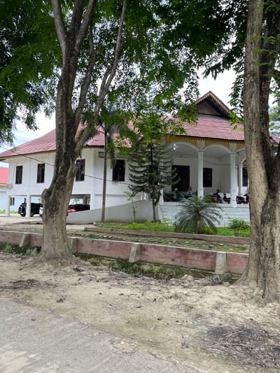 A side view of a white building with a red tiled roof and a covered porch is visible. The building appears to be on stilts. Several motorcycles are parked under the porch. A small palm tree and other greenery are located in front of the porch. The building is framed by the trunks and branches of two large trees in the foreground. The ground in front of the building is dirt and gravel, with a concrete path in the lower left corner. A low, red-bricked wall with concrete supports runs along the middle ground. The scene is likely in Bundar, Indonesia, given the architectural style and vegetation.