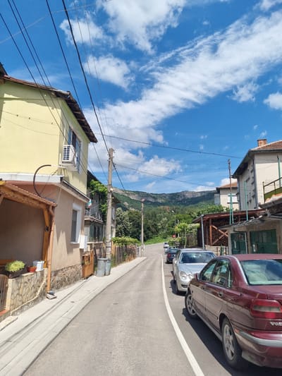 This image captures a street scene in the city of Belogradchik, Bulgaria, under a partly cloudy blue sky. A paved road with a white lane marking runs diagonally through the frame, curving to the right. On the left side of the road, a two-story building with a pale yellow facade and a brown tiled roof is visible. This building features windows, an air conditioning unit mounted near a window, and a covered porch area with wooden beams. Further down the street on the left, a Bulgarian flag is displayed on a balcony.
On the right side of the road, a series of buildings are present. The closest building has a light-colored facade, a red tiled roof with a chimney, and an attached structure with green doors. Next to this is another building with a more weathered facade and a covered area piled with firewood. Beyond that, additional houses and fences line the street.
Several cars are parked along the right side of the road. Closest to the viewer is a maroon Volvo sedan. Further down the road, a silver sedan, a black sedan, and a white car are parked in a row. In the background, the road continues, flanked by vegetation and leading towards a scenic mountain range with a rugged, rocky face and lush green slopes. Utility poles with numerous wires are prominent throughout the scene, indicating active infrastructure. The overall impression is of a residential street in a town situated in a hilly or mountainous region.