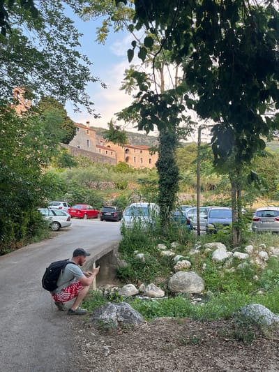 The image depicts a man sitting on a road in Podostrog, Montenegro, looking at his phone. He is wearing a grey t-shirt, red patterned shorts, a backpack, and a baseball cap. The road appears to be a rural street, with several cars parked along its side. In the background, a stone building with arched windows and a tiled roof is visible, surrounded by lush greenery and trees. The scene suggests a sunny day, possibly late afternoon, given the soft light and shadows. The overall atmosphere is relaxed and informal.