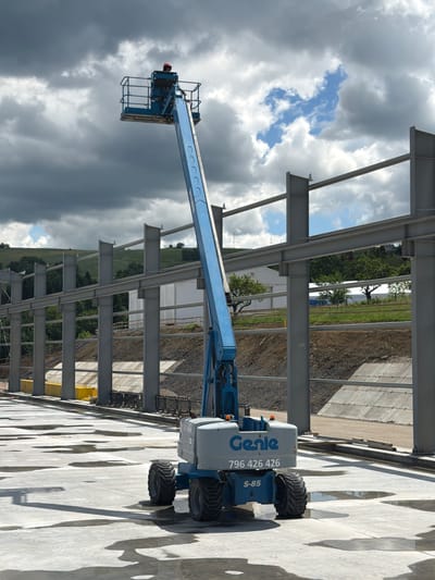 An aerial work platform, identified by the branding "Genie" and model number "S-85", is positioned on a wet, concrete surface. The platform is extended upwards and features a blue basket at the top, where a person wearing a red hard hat is visible. In the background, construction of a large steel-framed building is underway, with massive grey steel beams and columns forming the skeletal structure. The ground is a mix of excavated earth and concrete retaining walls. The environment appears to be a construction site located in Stróża, Poland, under a dramatic sky with dark, heavy clouds interspersed with patches of blue.