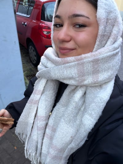 A female subject, approximately 20-30 years old, is depicted in a close-up, outdoors in Bouxwiller, France. She is wearing a cream-colored, coarsely woven scarf with faint pink stripe patterns, wrapped around her head and neck. A dark-colored padded jacket covers her torso. Her left hand holds a piece of toasted bread or crust. The subject is looking forward, directly at the camera, with a slight smile. In the background, behind the subject and to her left, a red two-door hatchback vehicle is parked on a paved surface, partially obscured by a light-colored building wall. The vehicle's roof and hood show a light dusting of snow. The overall environment appears to be an urban or residential setting.