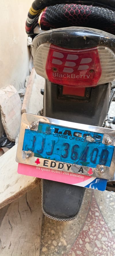 The image captures a close-up view of the rear section of a motorcycle or scooter, possibly parked in an outdoor setting in Kaura Goje, Nigeria. The primary subject is the vehicle's tail light assembly and license plate.
The tail light features a red lens with a prominent "BlackBerry" logo, consisting of the triple B symbol and the word "BlackBerry" beneath it. Below the tail light, a blue license plate is attached within a metallic frame. Visible text on the plate includes "LAGOS" (partially obscured) and "CENTRE OF EXCELLENCE" at the top, and the alphanumeric sequence "JJJ-36400D" centrally. Below the license plate, a pink and blue card or sticker is partially visible, bearing the text "EDDY A" next to a red clover-like symbol, followed by "the system ISO9001:2000 attestation".
The motorcycle's rear fender is black plastic, and parts of the seat or luggage rack are visible above, covered with black netting that has a red mesh pattern. The background on the left shows a rough, light-colored wall or concrete structure, with some debris on the ground. The ground beneath the vehicle is a tiled surface with a mosaic pattern of reddish and beige tones. No activity is taking place, and the scene appears static, suggesting the vehicle is stationary. The wear and tear on the vehicle parts, particularly the faded license plate and scratches on the fender, indicate regular use.