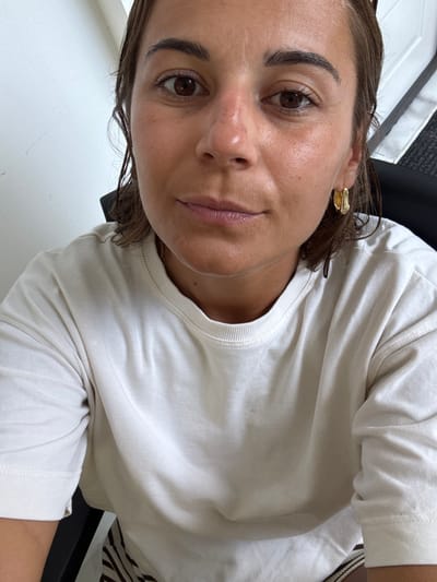 A woman looks at the camera with a slight smile. She has brown eyes and dark eyebrows. She wears a gold earring. Her hair is short and slightly damp. The woman is wearing a cream-colored t-shirt. The background includes a chair and a white wall. The lighting is soft and natural.