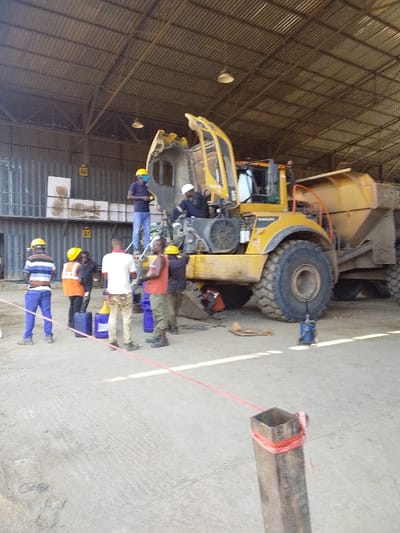The scene is set inside a large industrial shed with a corrugated metal roof and concrete floor, located in Hoche, Nigeria. A large yellow articulated dump truck, appearing to be a Volvo model, is undergoing maintenance. Its engine compartment covers are open, revealing internal components, and the front hood is raised.

Approximately ten adult males are present around the truck. Two individuals are positioned directly on or within the truck's engine bay: one man in a blue shirt, blue jeans, and a yellow hard hat stands on a platform near the engine, while another in dark coveralls and a white hard hat works deeper inside the compartment. A third individual in dark clothing and a white hard hat is partially visible, crouched beneath the front of the truck.

Other workers stand on the concrete floor around the vehicle. These include a man in a striped shirt, blue pants, and a yellow hard hat; a man in an orange safety vest and yellow hard hat; a man in a white t-shirt and light-colored pants; and a man in a red tank top, olive green pants, and a yellow hard hat. Most visible workers are wearing hard hats, and some have safety vests.

On the concrete floor, several blue plastic containers, possibly jerry cans, are visible. A blue hydraulic jack or similar tool is positioned near the front right wheel of the truck. Various tools and parts are scattered on the floor. A red rope, acting as a demarcation, extends from a metal post in the foreground towards the left side of the frame. The background shows corrugated metal walls, possibly with industrial shelving or platforms labeled "A4" and "A1". Hanging dome-shaped lights are visible on the ceiling.