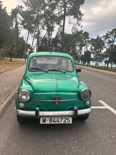 A vintage green Fiat 600 car is parked on the right side of a paved road. The car is facing forward and its front is visible. The car has two headlights, a chrome bumper, and a license plate that reads "M-844725". The car is on the Isle of Toxa, Spain, as indicated by the license plate prefix "M" which refers to the province of Madrid. The road is bordered by a gravel path on the left and a fence with trees behind it on the right. The sky is cloudy.