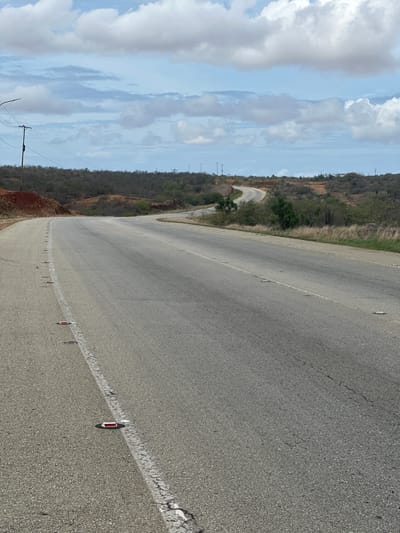 An asphalt road curves through a dry, hilly landscape under a partly cloudy sky. The road is marked with a white line on the left edge and has red reflectors intermittently placed along this line and further into the road. Power lines are visible on poles in the distance, and sparse vegetation, including cacti, dots the hillsides. The scene appears to be a rural or semi-rural area outside of a city.