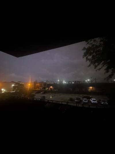This image, taken in Des Plaines, United States, depicts a dark, overcast sky, likely at dusk or during a storm. The foreground is dominated by a dark, silhouetted structure, possibly a balcony or roof overhang. Beyond this, a street or parking lot is visible with several cars parked. The background reveals buildings and what appear to be industrial structures, illuminated by various lights. A lone streetlamp casts an orange glow. The overall atmosphere is somber and subdued due to the low light conditions.