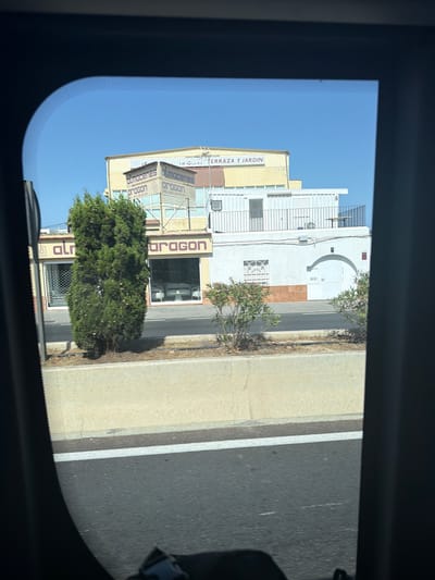 The image is taken through a car window, showing a view of buildings and a tree in the city of Santa Eulària des Riu, Spain. In the background, there is a light yellow building with "ALMACENES ARAGON" and "TERRAZA Y JARDIN" signs, suggesting it's a warehouse or store for garden supplies. To the left of this building, there's a storefront with "almacenes aragon" text on it, featuring large glass windows displaying what appears to be outdoor furniture. A tall, dense green tree stands in front of this storefront. To the right of the yellow building is a white structure with a balcony and railings. The foreground shows a concrete barrier and a road with a white line. The sky is clear and blue.