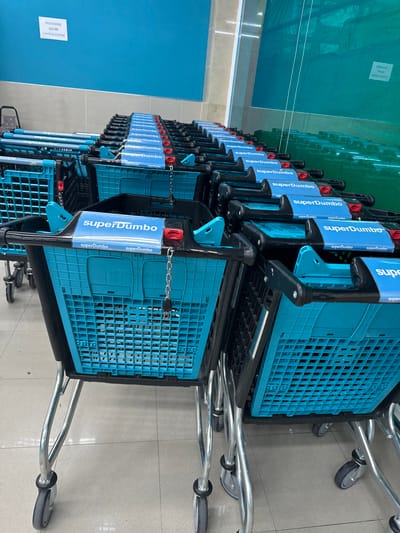 The image shows a row of blue shopping carts lined up in what appears to be a supermarket or similar retail environment. The carts are made of a plastic basket and a metal frame with wheels. Many of the carts have a blue sign with white lettering that reads "superDumbo" attached to the handle. The carts are positioned on a tiled floor, and in the background, there is a glimpse of a green mesh covering and a blue wall.
A white sign is affixed to the blue wall with text in black, which appears to be in Spanish. The text reads "PROHIBIDO SACAR CARROS-CESTAS", which translates to "PROHIBITED TO TAKE OUT CARTS". This suggests the carts are not meant to be removed from the premises.
The lighting suggests it is daytime, possibly indoors with artificial lighting. The overall scene is tidy and organized, typical of a retail setting ready for customers. There are no people visible in the image. The focus is entirely on the stacked shopping carts and the surrounding environment.