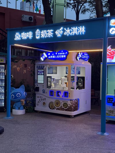 This outdoor shot, taken in the Nanshan District of China, features a robotic coffee kiosk. The kiosk is white, adorned with coffee bean images and signage in Chinese and English. The signage includes "AI机器人拉花咖啡," which translates to "AI Robot Latte Coffee" and "COFFEE." A blue robotic figure stands next to the kiosk. The surrounding area is a city street, with trees, other kiosks, and buildings visible in the background.