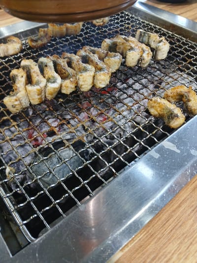 This is a close-up, overhead shot of unagi, or grilled eel, being cooked on a metal grate over hot coals. The eel pieces are arranged in several rows on the grate, some are curved and others are in straight segments. They appear to be glazed and are lightly charred in places, indicating they are actively cooking. The background shows the glowing red embers beneath the grate and a portion of the metallic grilling apparatus, along with a wooden surface on the right side, suggesting a restaurant setting. The image is likely taken in Suwon-si, South Korea, a city known for its culinary scene.