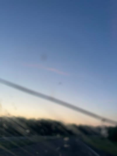 The image shows a blurred view of a landscape at dusk, possibly taken from a vehicle. The sky is a gradient of blue and pale yellow, with a faint pinkish hue near the horizon. A dark, diagonal line, possibly a power line or a reflection on a window, stretches across the upper portion of the image. Below this, a blurry line of trees or foliage is visible, with a dark road or ground in the foreground. The overall impression is one of a tranquil, transitional moment at the end of the day. The location in Krapivinskiy, Russia, is not visually discernible from the image itself; this information is external to the visual content.