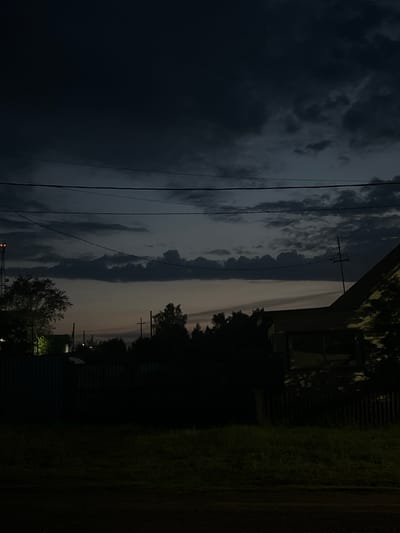 This image depicts a twilight scene with a dramatic sky. The sky is filled with dark, heavy clouds on the left, transitioning to lighter, wispy clouds towards the right. The scene appears to be outdoors, with power lines running horizontally across the frame. To the right, a dark silhouette of trees and a building with a fence are visible. The time of day is clearly evening or dusk, as indicated by the low light and the subdued colors. The weather appears to be overcast or possibly on the verge of rain, given the dark clouds. There are no people visible in the image. The setting seems to be a semi-rural or suburban area, with hints of man-made structures alongside natural elements like trees and grass. The overall mood is somber and atmospheric. No text is visible in the image.