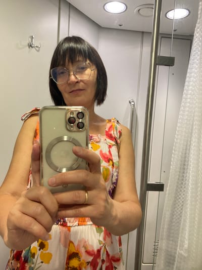 The image shows a woman taking a selfie in what appears to be a bathroom or changing room. She is wearing glasses and a floral-print dress. She is holding up a smartphone to take the photo. The background is a plain wall with some lighting fixtures and a metal bar. The overall setting suggests an indoor, possibly utilitarian space. The woman's expression is neutral. There is no visible text in the image. The lighting is bright and even. The rotation of the image suggests it was taken with the phone held vertically, and then rotated for viewing.