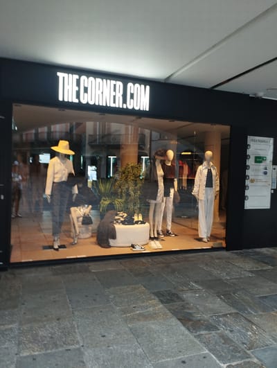 This image captures the storefront of "THE CORNER.COM", a fashion boutique located at Via Nassa 3 in Lugano, Switzerland. The shop features a large glass window displaying several mannequins dressed in contemporary clothing.
On the left side of the display, a mannequin is posed with a yellow hat, a white long-sleeved shirt, and dark trousers, holding a black hat and a black handbag. Another mannequin, partially visible and seated, is dressed in a white skirt and top, with a handbag.
To the right, two mannequins are displayed side-by-side. The first mannequin wears a black top, white trousers, and black sneakers. The second mannequin is dressed in a dark shirt and white trousers. Further to the right, a mannequin is presented in a white jacket and white trousers, with a dark top underneath.
In the background of the display, there is a large green plant and various clothing items are visible on racks inside the store. A sign on the right side of the storefront displays "VIA NASSA 3" and logos for "SOFAR" and "ZAFAR". The exterior pavement is made of large, grey stone slabs. The lighting suggests it is daytime, though possibly overcast or late in the day.