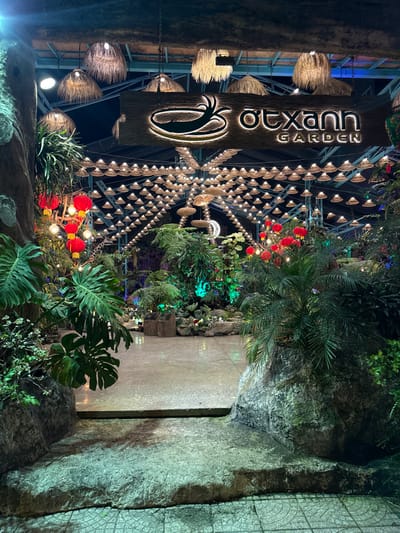 The image displays the illuminated nighttime entrance of a establishment named "ỚT XANH GARDEN" in An Hải Ward, Vietnam. A textured, light-colored concrete path forms the central entry point, originating from a tiled foreground and ascending a slight step. A prominent rectangular wooden sign, suspended overhead, features an illuminated logo depicting a stylized chili pepper within an oval, accompanied by the backlit text "ỚT XANH GARDEN". Above the pathway, an extensive ceiling arrangement consists of hundreds of small, conical, woven light fixtures, creating a grid-like canopy. Several larger, fringed straw light fixtures are also visible nearer the sign. Dense, lush tropical foliage, including large-leafed plants such as Monstera, ferns, and various smaller shrubs, frames both sides of the entrance path, some integrated into artificial rock formations. On the left, multiple clusters of red traditional lanterns are interspersed with string lights within the vegetation. Green and blue accent lighting illuminates specific plant areas further inside the garden, where a crescent moon-shaped light fixture is also visible. No individuals are present in the scene.