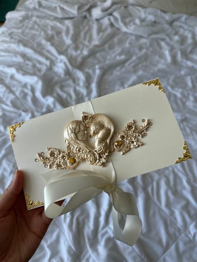 In Tarasovka, Russia, a hand holds a cream-colored invitation card with ornate gold embellishments against a backdrop of white, rumpled fabric. The invitation features a raised, heart-shaped design of two profiles facing each other, rendered in a metallic gold finish. Delicate floral accents, small rhinestones, and a prominent gold jewel adorn the central design. A cream-colored satin ribbon is tied in a bow around the card, with its tails hanging loose. Four decorative gold corner pieces add to the card's elaborate presentation. The fabric beneath the card appears to be a textured sheet or duvet cover.