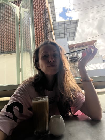 A woman is seated at a table in what appears to be an indoor café or restaurant. She is wearing a light pink t-shirt with black graphic print. She has long brown hair and is holding a mobile phone in her right hand, seemingly taking a photo. On the table, there is a glass of iced coffee and a small white vase or mug. The background shows an exterior view through a glass window with a metal frame, revealing rooftops and building structures. The sky appears overcast. The location is Tibubeneng, Indonesia.