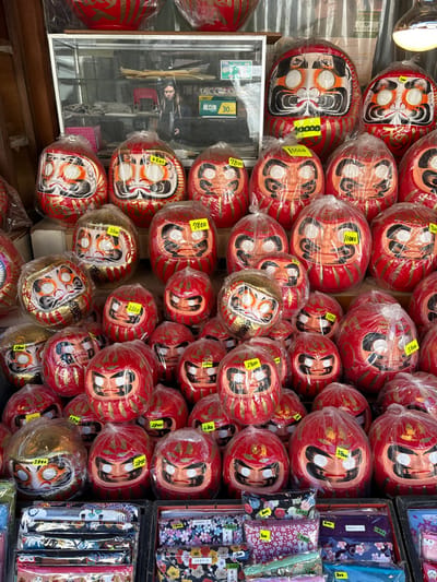 The image shows a display of traditional Japanese "daruma" dolls, located in a shop in Katsushika City, Japan. The dolls are red and have a white face with black features. They are wrapped in clear plastic and stacked in layers. Some have yellow stickers with prices on them. Behind the dolls, a clear glass display case shows items with Japanese text on them. A person, likely the store owner, is partially visible through the glass. The display case also has shelves with various colorful cloths, and a stack of books on a bottom shelf. The daruma dolls are a popular Japanese good luck charm. The scene appears to be in a shop or a street market. The environment is clean and well-lit.