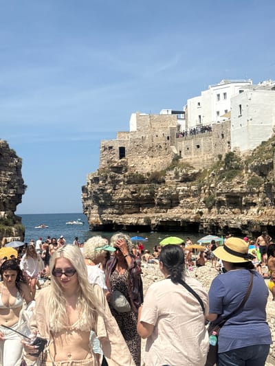 The image depicts a crowded beach scene in Polignano a Mare, Italy. White buildings and stone cliffs form the backdrop to the beach. Numerous people are visible, some sunbathing, others in the water, and some walking along the cliffs. Boats are seen on the water. The sky is clear and blue.