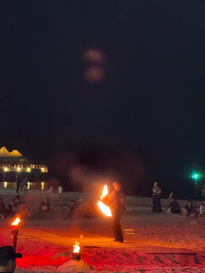 A nighttime scene on Cenang Beach, Malaysia, shows a fire performance. A single individual, wearing dark clothing, stands center-right, actively spinning a fire prop that creates bright orange, blurred arcs of flame. Smoke emanates from the performance. Two smaller, stationary fire pots are placed on the sandy ground in the foreground, casting a red-orange glow on the immediate area. Multiple spectators are visible across the sand, some seated, some standing, observing the performance. In the background, illuminated structures with light-colored, peaked roofs are visible near a body of water that reflects ambient lights. A distinct green light source appears on the far right in the distance. The sky above is dark.